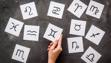 Mão segurando um cartão com símbolo de Gêmeos, cercada por outros cartões com signos do zodíaco sobre fundo cinza.