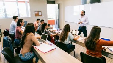 Imagem de uma sala de aula com alguns alunos sentados um ao lado do outro e um professor à frente do quadro branco, ministrando a sua aula.