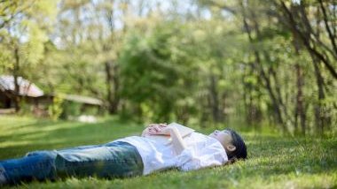 Imagem de uma mulher oriental deitada em um gramado embaixo de uma árvore relaxando. Sobre o seu peito, um livro aberto.