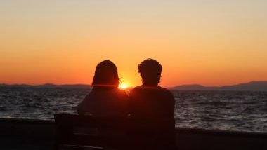 Imagem de um casal sentado em um banco olhando para o mar. Ao fundo, um lindo pôr do Sol.