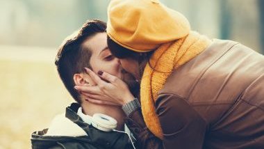 Imagem de um casal usando roupas de inverno, namorando e se beijando em uma praça.