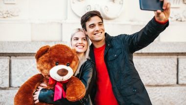 Imagem de um casal apaixonado, fazendo um selfie. Ela está segurando um lindo e gigante urso de pelúcia com um laço vermelho no pescoço.
