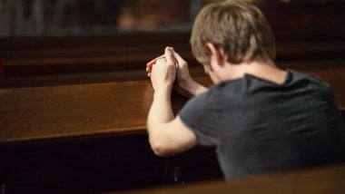 Imagem de um homem dentro de uma igreja ajoelhado fazendo a sua oração em um banco de madeira.