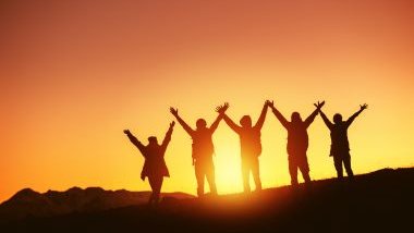 Imagem da silhueta de várias pessoas felizes, posicionadas sobre uma montanha, com as mãos para o alto e ao fundo, o pôr do Sol.
