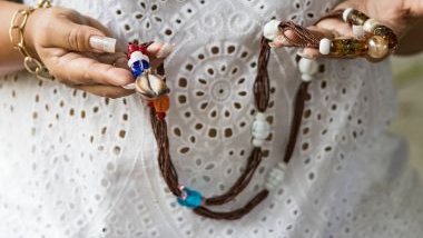 Imagem de uma mãe de santo, usando um vestido de laise branco, segurando em suas mãos uma guia com conchas e pedras coloridas.