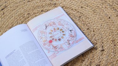 Imagem de um livro aberto sobre uma toalha feita de palha. Na página, uma imagem com uma mandala astrológica e os 12 signos do zodíaco.