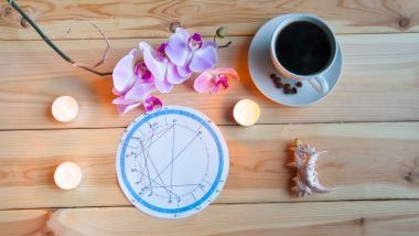 Imagem de uma mesa de madeira e sobre ela uma mandala astrológica, uma xícara de café, uma haste de orquídea e três velas. 