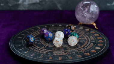 No foco da imagem, há uma Roda do Zodíaco com os símbolos dos signos e há dados em cima dela. Ao lado, há uma bola de cristal.