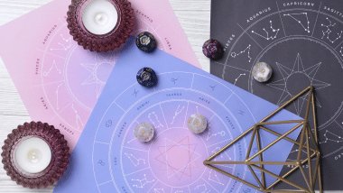 Há três rodas do zodíaco com os símbolos dos signos desenhadas em papel sulfite colorida em cima de uma mesa. Ao redor, há velas e dados.