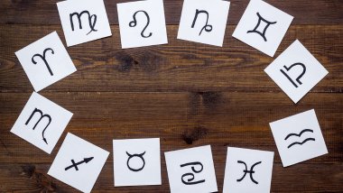 Cada símbolo de um signo do zodíaco está desenhado em um pedaço de papel. Os papéis estão espalhados em cima de uma mesa de madeira.