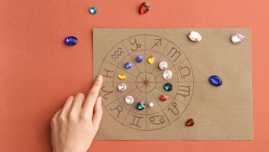 Há uma roda do zodíaco com os símbolos dos signos desenhada em papel cartão em cima de uma mesa. Ao redor, há pedras.