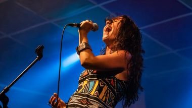 Imagem de uma mulher morena, cantora, em cima de um palco, segurando o microfone.