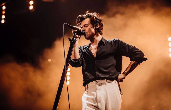 Harry Styles segurando microfone com uma mão, e a outra mão em sua cintura, durante show.