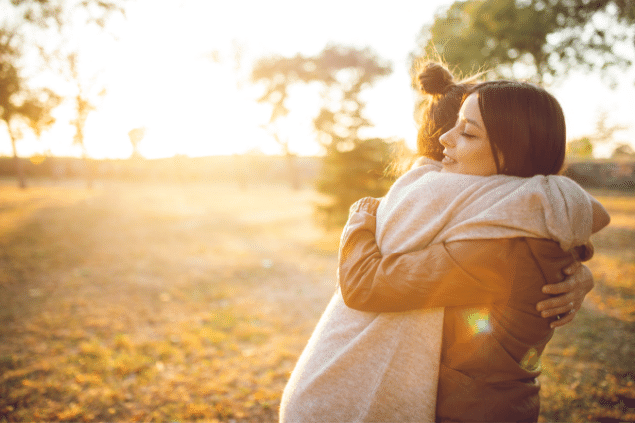 Duas mulheres se abraçando em um quintal cheio de folhas secas, ao pôr do sol.