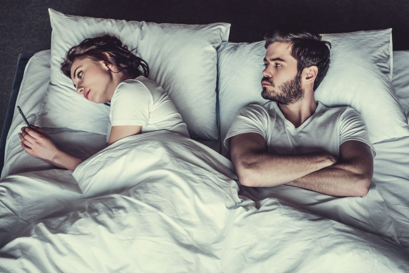 Há um casal deitado em uma cama. A mulher mexe em seu celular enquanto o homem tenta olhar o que ela está fazendo, com expressão séria.
