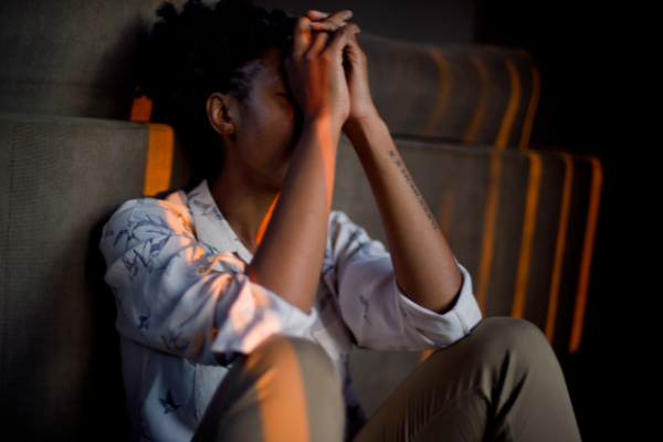 Mulher triste sentada no chão apoiando os dois cotovelos sob os joelhos enquanto protege o rosto com as mãos