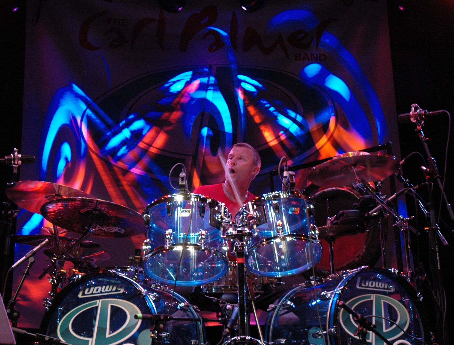 Carl Palmer tocando bateria no palco com luzes coloridas