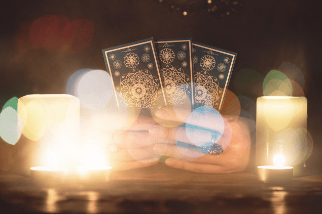 As mãos de uma taróloga segurando cartas de Tarot estão em destaque na imagem. Ao lado, há velas.