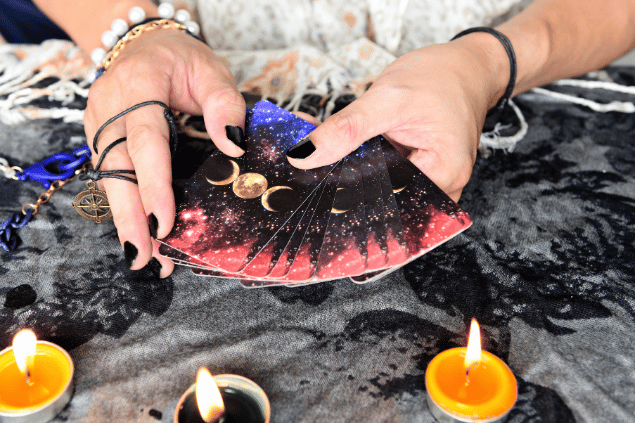 As mãos de uma taróloga segurando cartas de Tarot viradas para baixo estão em destaque na imagem. Ao lado, há velas e outros objetos.