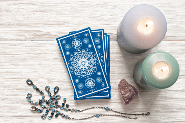 Algumas cartas de Tarot viradas para baixo estão em destaque na imagem. Ao lado, há duas velas e outros objetos.