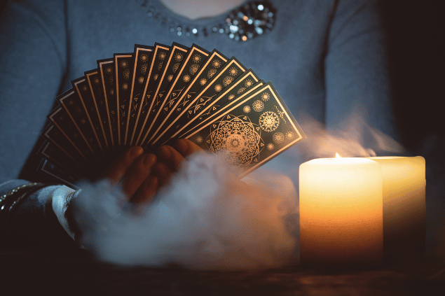 As mãos de uma taróloga segurando cartas de Tarot estão em destaque na imagem. Há duas velas ao lado.