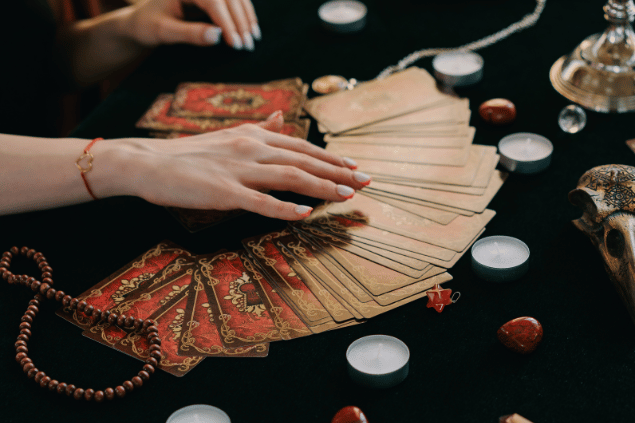 As mãos de uma taróloga posicionando cartas de Tarot viradas para baixo estão em destaque na imagem. Ao lado, há velas e pedras.