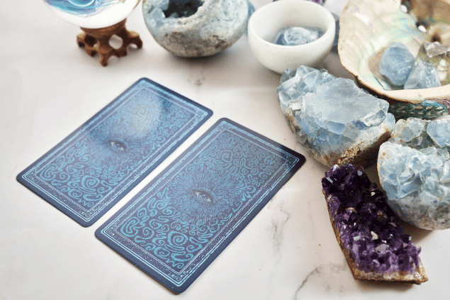 Duas cartas de Tarot viradas para baixo estão em destaque na imagem. Ao lado, há alguns cristais e outros objetos.