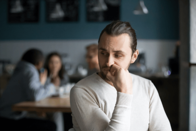 Homem chateado sentado sozinho em um café. Ele está longe do seu grupo de colegas. 