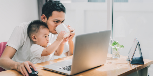 Homem sentado à mesa segura um bebê que coloca caneca próximo de seu rosto. O homem mexe em um computador.