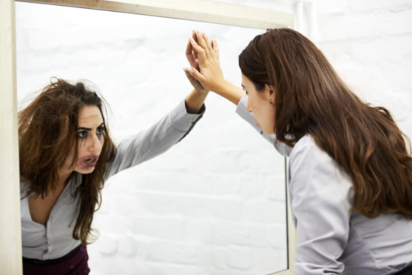 Mulher olhando seu reflexo no espelho. No reflexo, o cabelo está bagunçado e sua maquiagem borrada