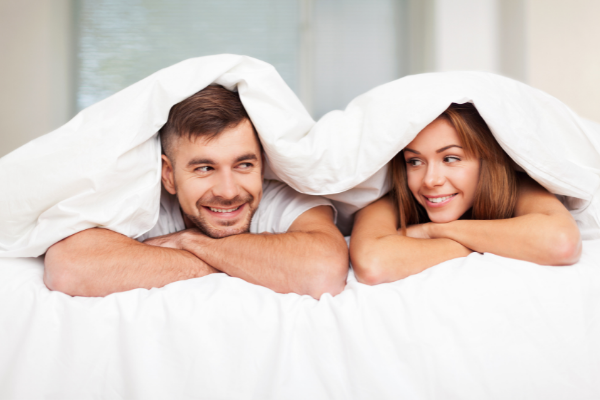 Homem e mulher debaixo de uma coberta. Os dois estão sorrindo