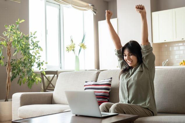 Imagem de uma mulher com as mãos erguidas e sorrindo sentada no sofá da sala e na frente do notebook