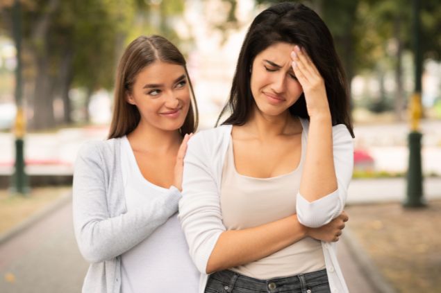 Imagem de uma mulher chorando e a outra atrás dela sorrindo, enquanto finge que a consola