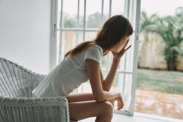 Imagem de uma mulher sentada em sua sala com a mão na cabeça como se estivesse triste