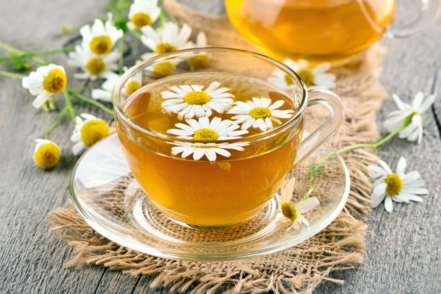 Imagem de um chá de camolila com as flores em volta em cima de uma mesa