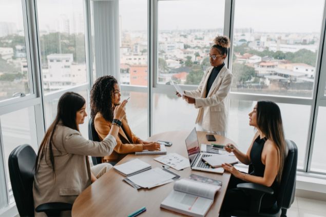 Imagem de uma mulher liderando uma reunião com mais três mulheres em uma sala de reuniões