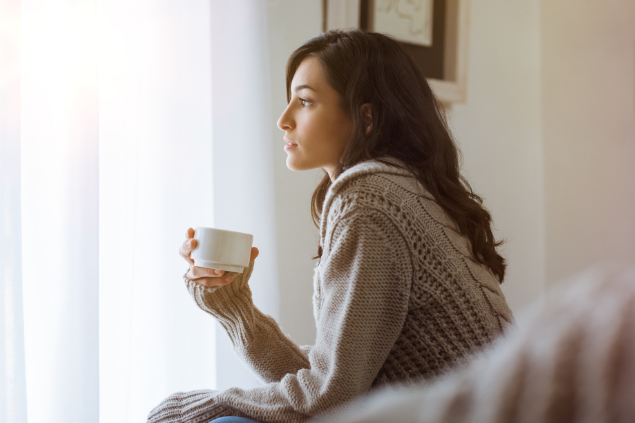 Mulher sentada com um copo de café na mão enquanto olha para fora da janela