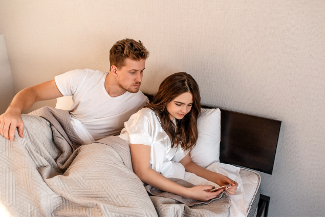 Casal deitado na cama enquanto o homem observa a mulher que mexe no celular