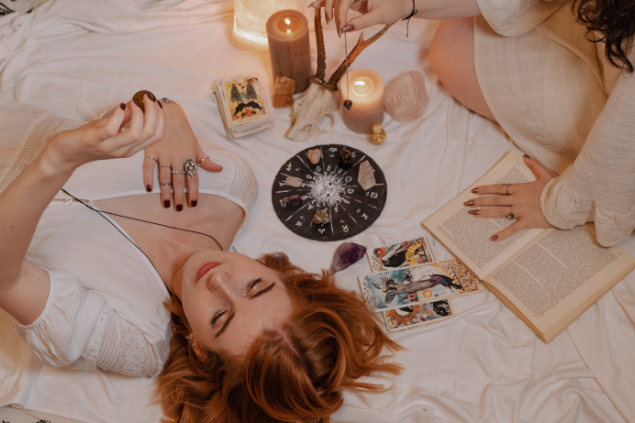 Mulher deitada ao chão segurando uma pedra e ao redor de pedras, cartas de tarot e outros objetos 