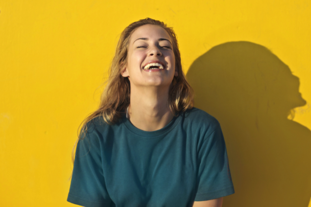 Mulher loira sorrindo e ao fundo, uma parede amarela com cor bem vibrante