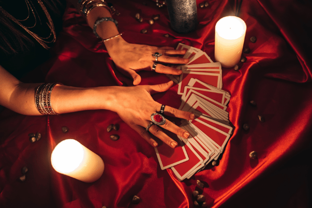 Uma vidente ou taróloga tem as suas mãos no centro da imagem. Ela as coloca sobre as cartas de tarot dispostas na mesa.