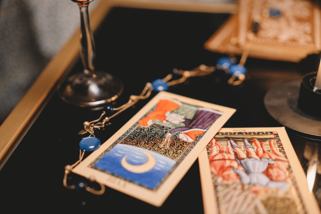 Duas cartas de Tarot estão sobre uma mesa no foco da imagem. Ao redor delas, há outros objetos.