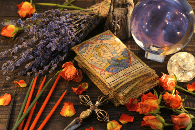 Um baralho de cartas de Tarot está sobre uma mesa no foco da imagem. Ao redor dele, há uma bola de cristal, flores, pétalas e outros objetos.