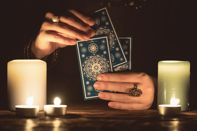 As duas mãos de uma mulher estão segurando três cartas de tarot. Ao lado, há cinco velas.