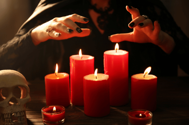 Uma mulher que veste um capuz preto está com as suas duas mãos no ar e por cima de sete velas acesas.
