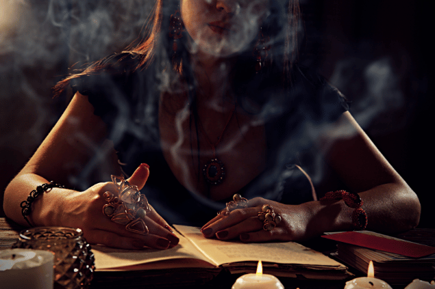 Uma mulher está lendo um livro que está sobre uma mesa. Na mesa, há outros itens como velas e baralhos de tarot. Ao redor dela, sobe uma fumaça.