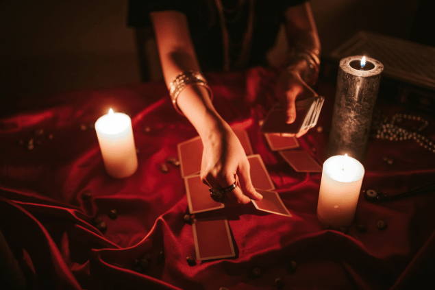 As mãos de uma taróloga segurando cartas de Tarot viradas para baixo estão em destaque na imagem. Ao lado e em cima de uma mesa, há velas e pedras.
