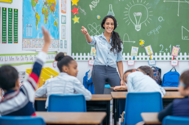 Uma professora está dando aula. Ela sorri e um dos alunos levanta a mão para falar.
