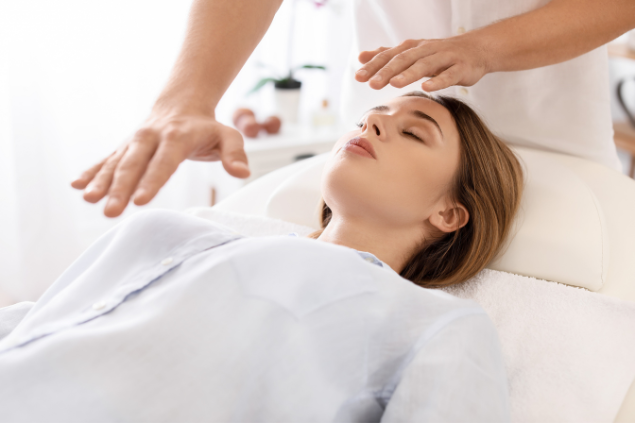 Imagem de uma mulher deitada sobre uma maca, usando roupas brancas. Ela está recebendo uma aplicação de reiki.