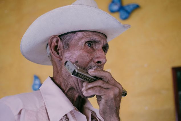 Imagem de um senhor usando um chapéu bege, tocando gaita. A foto traz o simbolismo do Pai Benedito na Umbanda.